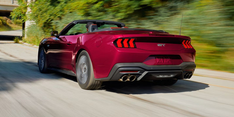 Red Ford Mustang convertible driving on a road with motion blur and greenery in the background.