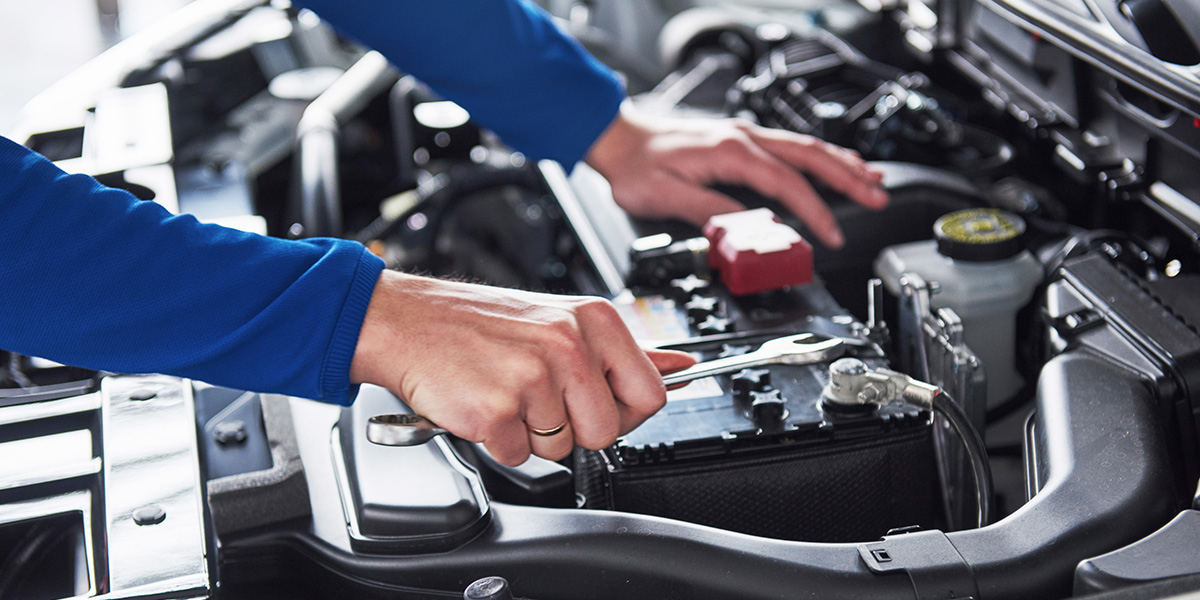 A mechanic working on a car battery with a wrench under the open hood of a vehicle.
